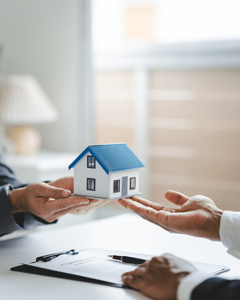 Twee handen geven een maquette van een huis door boven een contract